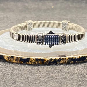 May include: A silver bracelet with a ribbed design and a dark blue, gem-studded clasp. The bracelet is displayed on a decorative, round agate stone with a brown and gold edge. The background is a neutral grey surface.