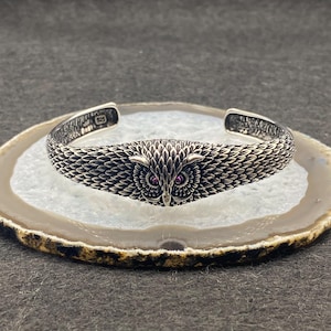 May include: A silver cuff bracelet featuring an owl design. The owl's face is the focal point, with intricate feather detailing and two small, round, pink gemstones for eyes. The bracelet is likely made of sterling silver, as indicated by the "925" stamp.