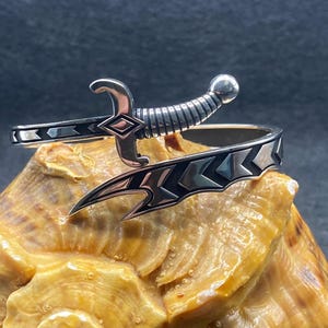 Puede incluir: Una pulsera de puño plateada con un diseño de espada. La pulsera presenta una empuñadura y una hoja detalladas, con patrones geométricos a lo largo de la banda. La espada está colocada en la parte superior de la pulsera, agregando un estilo único y atrevido.
