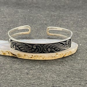 May include: A silver cuff bracelet with an engraved black band featuring a swirling, floral design. The open-ended bracelet is displayed on a light-colored, patterned surface against a dark gray background.