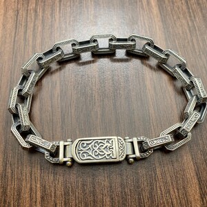 May include: Silver-toned bracelet with a rectangular link design. The links are connected and feature intricate engraved patterns. The clasp has a decorative design. The bracelet is displayed on a wooden surface.