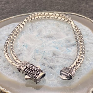 May include: A silver bracelet with a double-strand woven design. The clasp is rectangular and adorned with small, dark stones. The bracelet is displayed on a light-colored, marbled surface. The bracelet is a piece of jewelry.