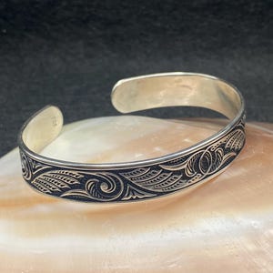 May include: Silver cuff bracelet with intricate, dark etched floral and swirl patterns. The open design allows for easy wear. The bracelet is displayed on a light-colored, textured surface.