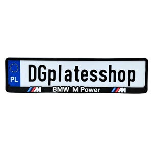 Op de afbeelding: Rechthoekige nummerplaat met de tekst "DGplatesshop" in zwart. De plaat heeft een witte achtergrond met een zwarte rand. De onderkant van de plaat leest "BMW M Power" met het BMW-logo aan elke kant. De letters "PL" en een blauwe cirkel met gele sterren staan links.