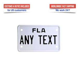 Può includere: Targa bianca con la scritta "FLA" in alto e "ANY TEXT" in grassetto nero in basso. La targa ha quattro fori, uno in ogni angolo. Il testo "CUSTOMS & DUTIES INCLUDED for US customers" è sopra.