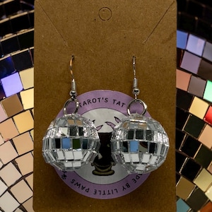 May include: A pair of silver disco ball earrings with a mirrored surface, suspended from silver hooks. The earrings are presented on a brown card with a purple sticker reading "Tarot's Tat" and "Made by Little Paws". The background is a disco ball.