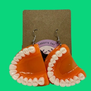 May include: Novelty earrings in the shape of orange dentures with white teeth, suspended from silver hooks. The earrings are displayed against a brown card with the text "TAROT'S TAT" on a purple background, set against a green backdrop.