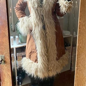 May include: A brown coat with a faux fur trim around the collar, cuffs, and down the front. The coat has a long, flowing design and is worn with black boots. The coat appears to be made of a soft, suede-like material.
