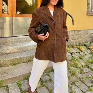 May include: A brown suede blazer with a classic collar and button closure. The blazer is paired with white trousers and a small black handbag. The outfit is completed with brown shoes. The blazer has two front pockets.