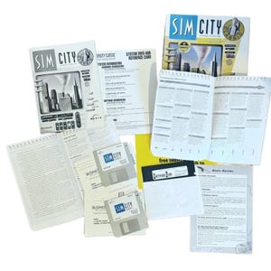 May include: A collection of vintage SimCity computer game materials, including the game box, manuals, floppy disks, and reference cards. The box features a cityscape graphic with the game title. The materials are scattered on a white surface.