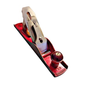 May include: A vintage red and silver hand plane with a wooden handle. The plane has a metal blade and is marked "CORSAIR U.S.A." on the side. This woodworking tool is designed for shaping and smoothing wood surfaces.