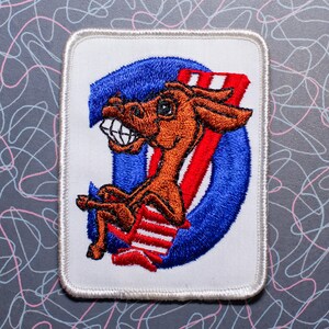 May include: Embroidered patch featuring a cartoon donkey in a red and white striped chair, set against a blue circle and red stripes. The donkey has a wide grin and is brown. The patch has a white border.