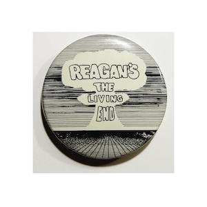 May include: A round button with a black and white striped background. The button features the text "REAGAN'S THE LIVING END" in a white cloud-shaped design. The design includes a mushroom cloud graphic.
