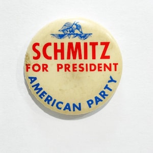 Könnte beinhalten: Ein cremefarbener Vintage-Button mit dem Text "SCHMITZ FOR PRESIDENT AMERICAN PARTY" in Rot und Blau. Der Button zeigt oben ein blaues Adlerdesign. Ein politischer Wahlkampf-Button.