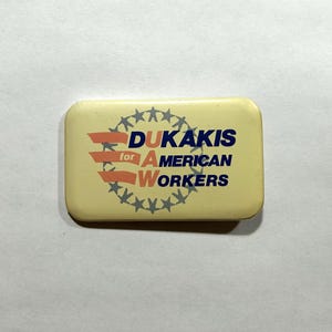 May include: A rectangular button with the text "DUKAKIS for AMERICAN WORKERS" in blue and red. The button has a light yellow background with a ring of grey stars around the text.