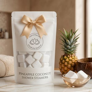 May include: A white bag with a gold bow contains white shower steamers. The bag has a clear window and text that reads "Pineapple Coconut Shower Steamers." A pineapple, coconut bowl, and dried pineapple slices are also visible.