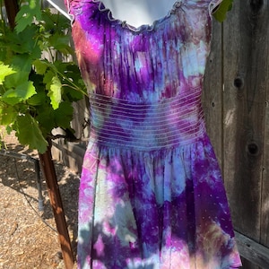 Puede incluir: Un vestido sin mangas con un vibrante estampado tie-dye en tonos morado, rosa y azul. El vestido presenta un escote con volantes y una cintura fruncida. La falda tiene un dise&ntilde;o en capas, y el vestido se exhibe en un maniqu&iacute;.