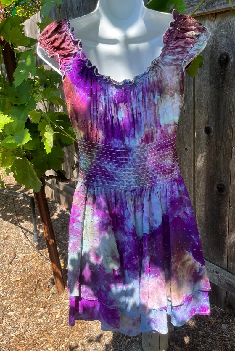 Puede incluir: Un vestido sin mangas con un vibrante estampado tie-dye en tonos morado, rosa y azul. El vestido presenta un escote con volantes y una cintura fruncida. La falda tiene un dise&ntilde;o en capas, y el vestido se exhibe en un maniqu&iacute;.