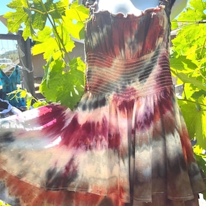 K&ouml;nnte beinhalten: Ein Batik-Kleid mit flie&szlig;endem Rock und R&uuml;schentr&auml;gern. Das Kleid hat eine Mischung aus Farben, darunter Rot, Lila und Braun, mit einem gepunkteten Muster auf dem Rock. Das Kleid wird im Freien auf einer Schaufensterpuppe pr&auml;sentiert.