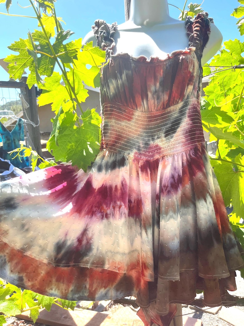 K&ouml;nnte beinhalten: Ein Batik-Kleid mit flie&szlig;endem Rock und R&uuml;schentr&auml;gern. Das Kleid hat eine Mischung aus Farben, darunter Rot, Lila und Braun, mit einem gepunkteten Muster auf dem Rock. Das Kleid wird im Freien auf einer Schaufensterpuppe pr&auml;sentiert.