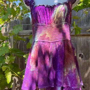 Puede incluir: Un vibrante vestido morado con falda escalonada y corpi&ntilde;o ajustado. El vestido presenta un escote cuadrado y mangas cortas. La tela tiene un patr&oacute;n texturizado de puntos y un efecto tie-dye con tonos de rosa, amarillo y morado. El vestido se exhibe en un maniqu&iacute;.