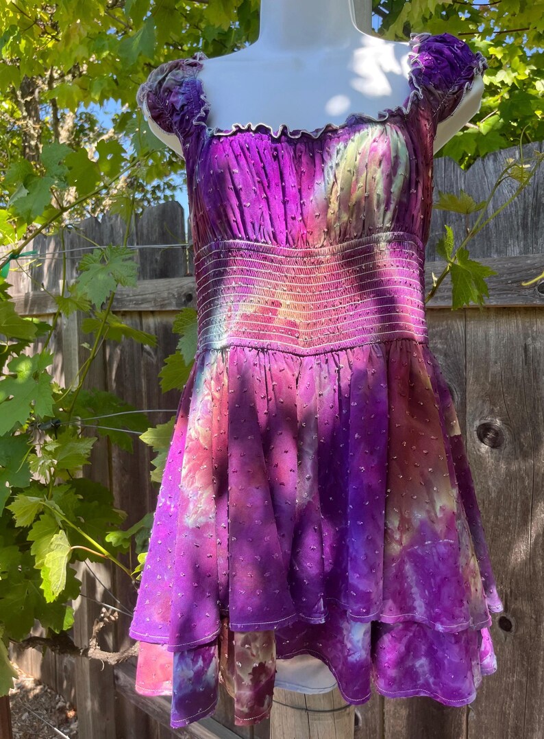 Puede incluir: Un vibrante vestido morado con falda escalonada y corpi&ntilde;o ajustado. El vestido presenta un escote cuadrado y mangas cortas. La tela tiene un patr&oacute;n texturizado de puntos y un efecto tie-dye con tonos de rosa, amarillo y morado. El vestido se exhibe en un maniqu&iacute;.