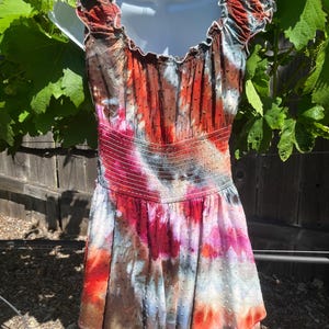 K&ouml;nnte beinhalten: Ein farbenfrohes Batik-Kleid mit ger&uuml;schtem Ausschnitt und taillierter Taille. Das Kleid zeigt eine Mischung aus roten, orangen, rosa und blauen Farbt&ouml;nen, die ein lebendiges und k&uuml;nstlerisches Design ergeben. Der Stoff hat eine subtile gepunktete Textur.