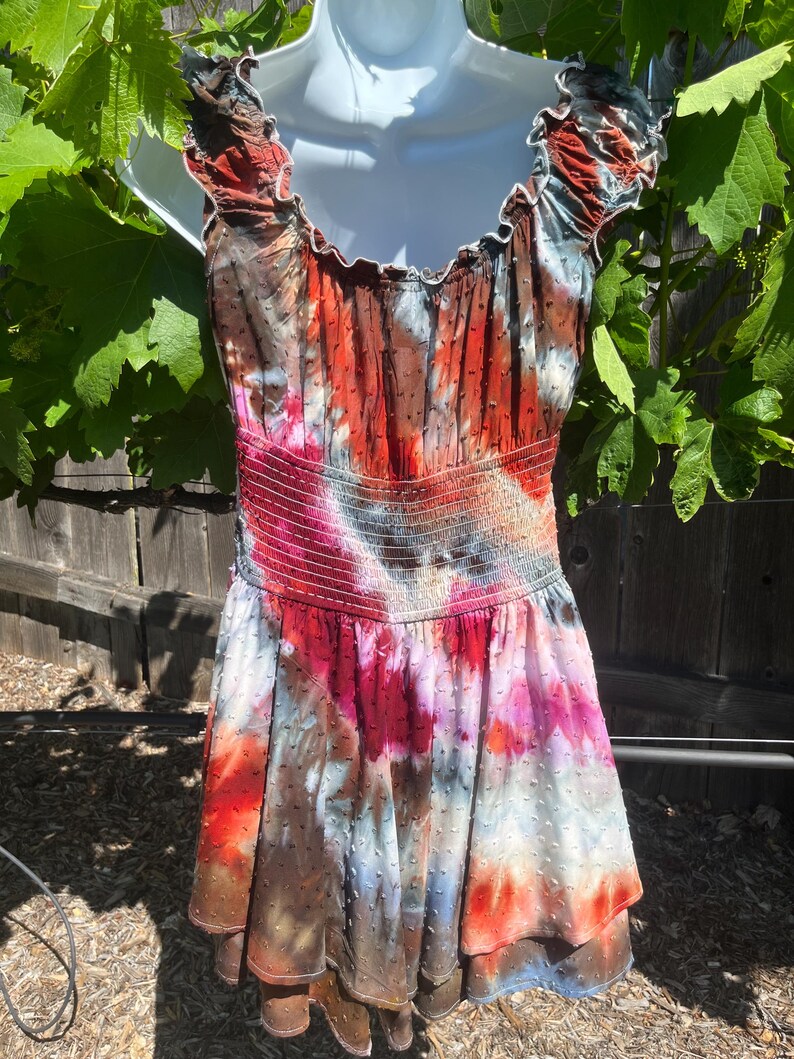 K&ouml;nnte beinhalten: Ein farbenfrohes Batik-Kleid mit ger&uuml;schtem Ausschnitt und taillierter Taille. Das Kleid zeigt eine Mischung aus roten, orangen, rosa und blauen Farbt&ouml;nen, die ein lebendiges und k&uuml;nstlerisches Design ergeben. Der Stoff hat eine subtile gepunktete Textur.