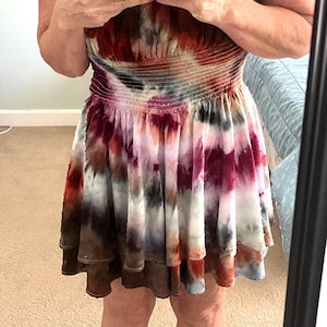 K&ouml;nnte beinhalten: Ein Mini-Kleid mit Batikmuster und R&uuml;schenausschnitt und -rock. Das Kleid hat eine Mischung aus Farben, darunter Rot, Wei&szlig;, Braun und Blau. Die Person tr&auml;gt schwarze Flip-Flops.