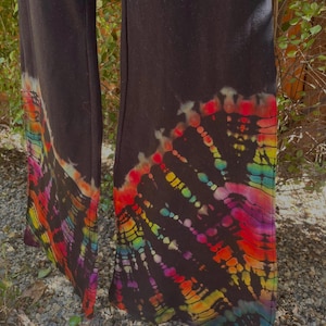 May include: Black pants with a vibrant tie-dye pattern at the hem. The tie-dye design features a spectrum of colors, including red, orange, yellow, green, blue, and purple, creating a bold and eye-catching look. The pants are made of a soft material.