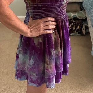Puede incluir: Un minivestido de terciopelo en tonos morados, azules y marrones. El vestido presenta un corpi&ntilde;o ajustado con cintura ce&ntilde;ida y una falda escalonada. El escote y las mangas tienen un ribete con volantes.