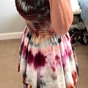 K&ouml;nnte beinhalten: Ein Batik-Kleid mit ger&uuml;schtem Ausschnitt und ausgestelltem Rock. Das Kleid hat eine Mischung aus Farben, darunter Burgunderrot, Rosa und Wei&szlig;. Die Person tr&auml;gt schwarze Flip-Flops.