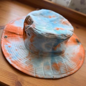 May include: A bucket hat featuring a tie-dye pattern in shades of light blue and orange. The hat has a wide, circular brim with visible stitching and small metal eyelets on the sides for ventilation, creating a casual headwear accessory.