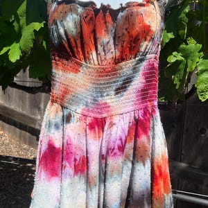 K&ouml;nnte beinhalten: Ein farbenfrohes Batik-Kleid mit einem gerafften Oberteil und R&uuml;schentr&auml;gern. Das Kleid zeigt eine Mischung aus roten, orangen, rosa und blauen Farbt&ouml;nen. Der Rock hat eine gepunktete Textur und einen gestuften Saum, was einen verspielten Look erzeugt.