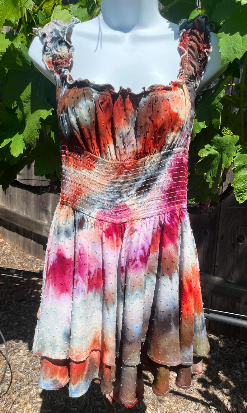 K&ouml;nnte beinhalten: Ein farbenfrohes Batik-Kleid mit einem gerafften Oberteil und R&uuml;schentr&auml;gern. Das Kleid zeigt eine Mischung aus roten, orangen, rosa und blauen Farbt&ouml;nen. Der Rock hat eine gepunktete Textur und einen gestuften Saum, was einen verspielten Look erzeugt.