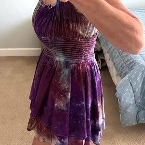 Puede incluir: Un vestido morado y azul te&ntilde;ido con un estampado tie-dye con una falda escalonada. El vestido tiene un corpi&ntilde;o ajustado con cintura fruncida y mangas cortas con volantes. La persona lleva chanclas negras.