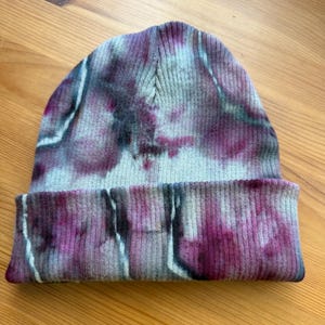 May include: A ribbed knit beanie featuring a tie-dye pattern with blended shades of deep magenta, soft grey, and white. The hat has a folded cuff at the base and a rounded crown, showcasing a textured fabric construction with a multi-colored effect.