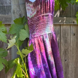 Puede incluir: Vestido te&ntilde;ido anudado en tonos morados, rosas y blancos, con una tela texturizada con lunares. El vestido presenta un cuello halter, una cintura fruncida ajustada y una falda escalonada. El vestido se exhibe al aire libre, colgado de una valla de madera.
