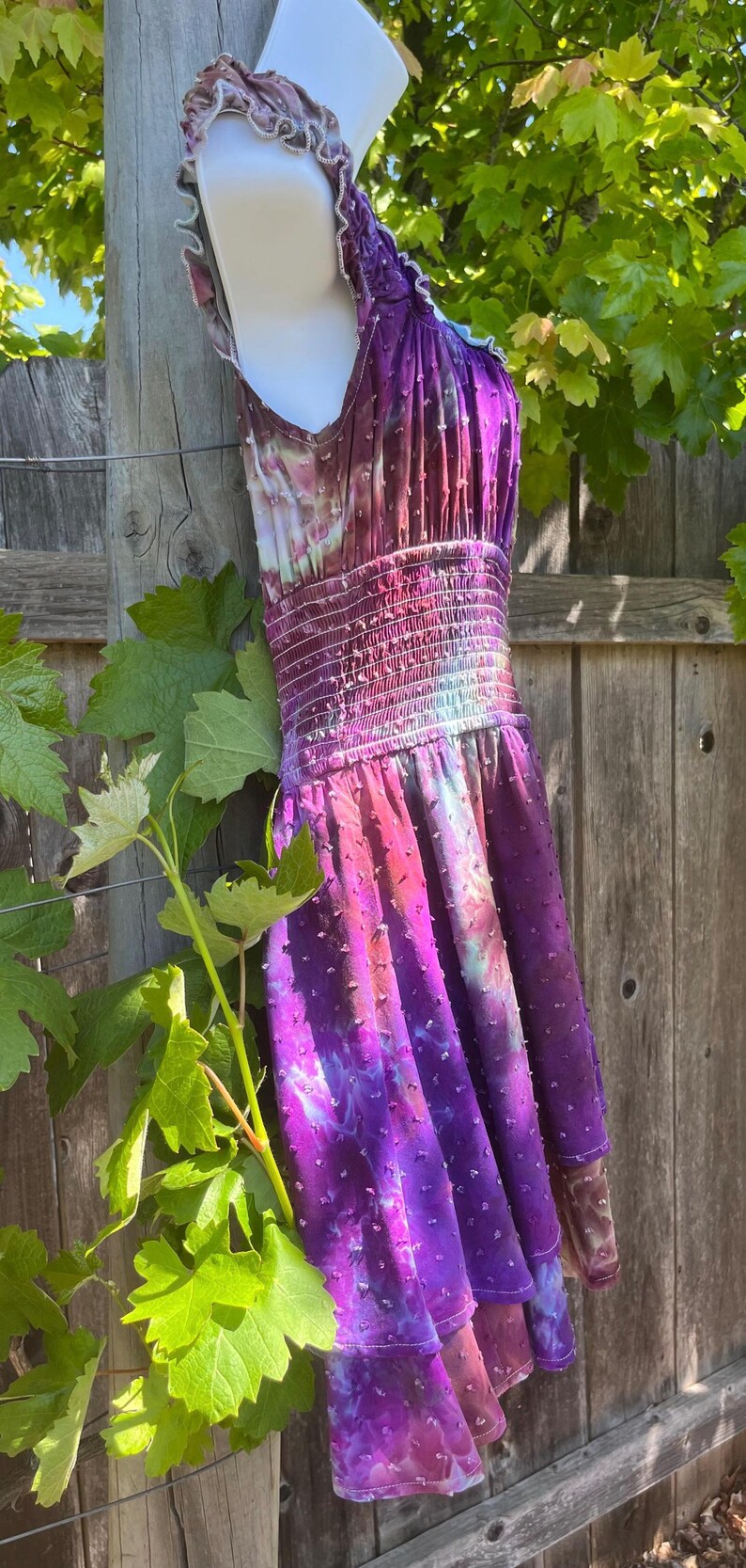Puede incluir: Vestido te&ntilde;ido anudado en tonos morados, rosas y blancos, con una tela texturizada con lunares. El vestido presenta un cuello halter, una cintura fruncida ajustada y una falda escalonada. El vestido se exhibe al aire libre, colgado de una valla de madera.