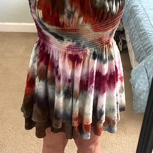 K&ouml;nnte beinhalten: Ein Mini-Kleid im Batik-Look in Rot-, Braun-, Wei&szlig;- und Lilat&ouml;nen. Das Kleid hat einen ger&uuml;schten Ausschnitt und ein gerafftes Oberteil. Der Rock ist gestuft und reicht bis zur Mitte des Oberschenkels. Die Person tr&auml;gt schwarze Flip-Flops.