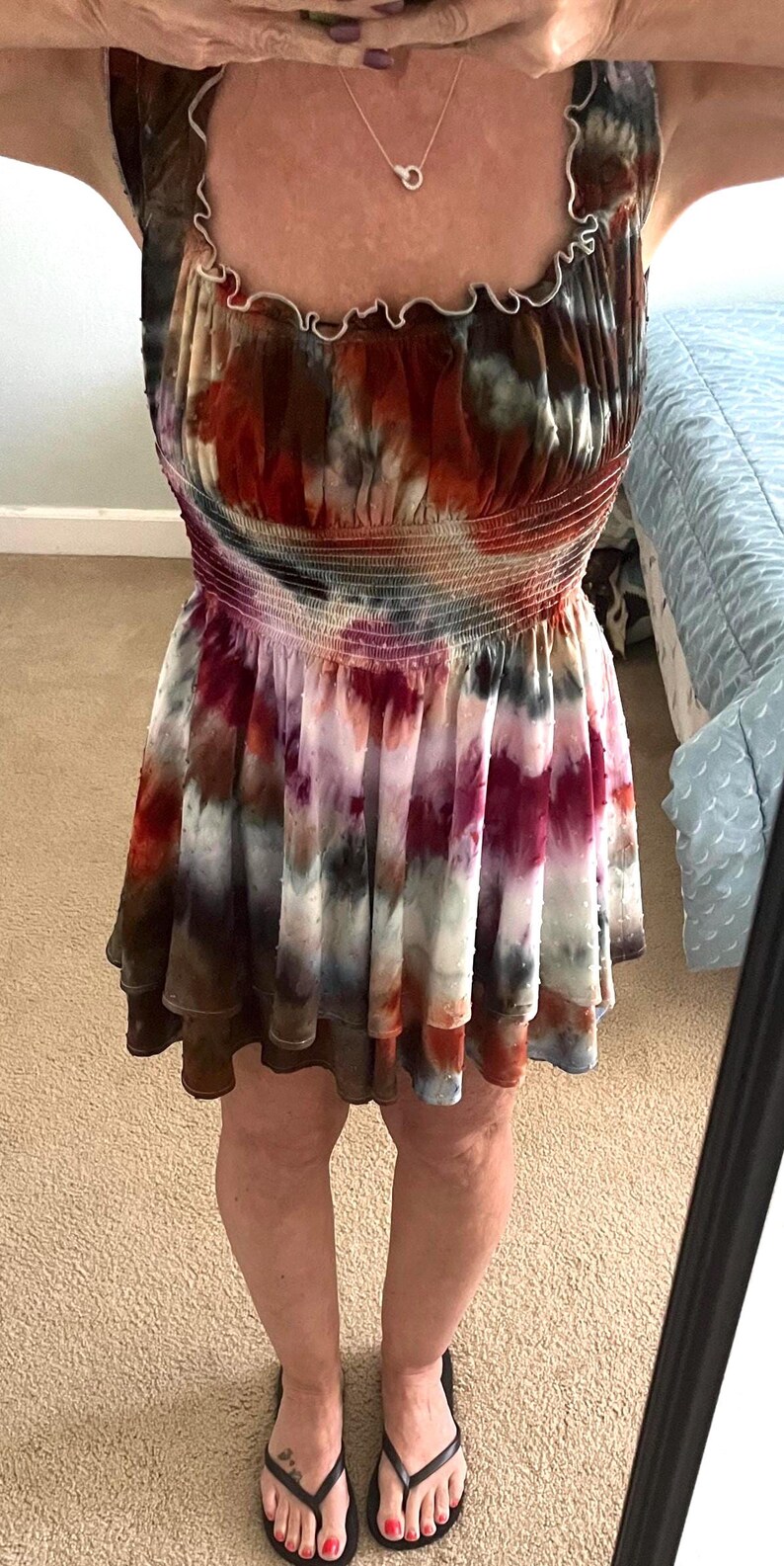 K&ouml;nnte beinhalten: Ein Mini-Kleid im Batik-Look in Rot-, Braun-, Wei&szlig;- und Lilat&ouml;nen. Das Kleid hat einen ger&uuml;schten Ausschnitt und ein gerafftes Oberteil. Der Rock ist gestuft und reicht bis zur Mitte des Oberschenkels. Die Person tr&auml;gt schwarze Flip-Flops.