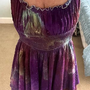 Puede incluir: Vestido morado sin mangas con cuello redondo y cintura entallada. El vestido presenta un estampado tie-dye con toques de verde y blanco. El dobladillo es con volantes y el vestido tiene una tela texturizada con lunares.