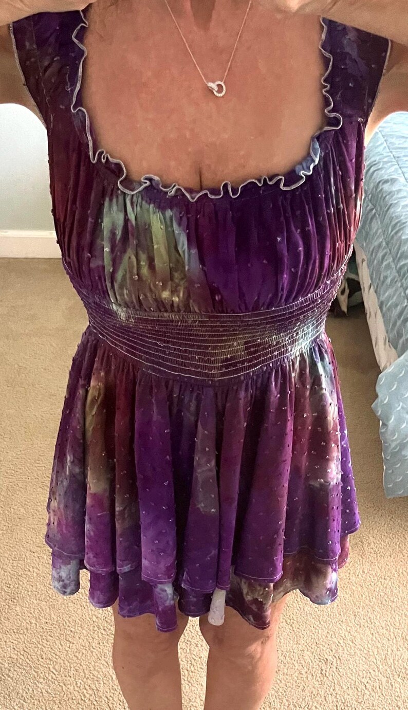 Puede incluir: Vestido morado sin mangas con cuello redondo y cintura entallada. El vestido presenta un estampado tie-dye con toques de verde y blanco. El dobladillo es con volantes y el vestido tiene una tela texturizada con lunares.