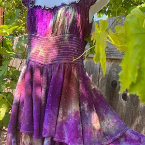 Puede incluir: Vestido morado vibrante con falda escalonada y corpi&ntilde;o ajustado. El vestido presenta un efecto tie-dye con tonos morados, rosas y blancos. Tiene un escote con volantes y mangas cortas. La tela tiene una textura punteada.