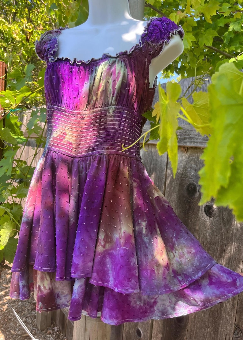Puede incluir: Vestido morado vibrante con falda escalonada y corpi&ntilde;o ajustado. El vestido presenta un efecto tie-dye con tonos morados, rosas y blancos. Tiene un escote con volantes y mangas cortas. La tela tiene una textura punteada.