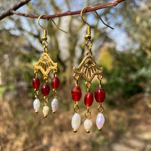 May include: A pair of gold-colored dangle earrings. Each earring features a decorative filigree top, red glass beads, and white pearl beads. The earrings are suspended from gold-colored ear wires.