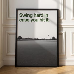 May include: Black framed print with the text "Swing hard in case you hit it." The image is a black and white photograph of a golf course with a flag on the green. The print is displayed against a white wall.