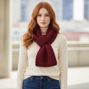 May include: A woman wearing a burgundy crochet scarf over a cream-colored turtleneck sweater and blue jeans. The scarf is wrapped around the neck and crossed in the front. The background is blurred, with a building visible.