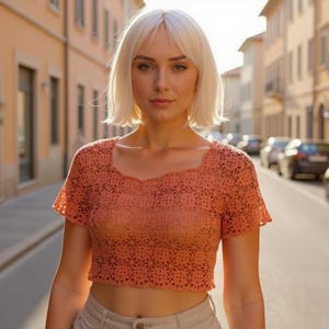 May include: A woman wearing a peach-colored, short-sleeved, crochet crop top. The top has a square neckline and a floral pattern. She is standing on a street with buildings and cars in the background.