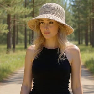 May include: A woman in a beige woven sun hat and a black sleeveless top stands on a dirt path in a forest. The hat has a wide brim and a rounded crown. The forest is filled with tall, green trees.
